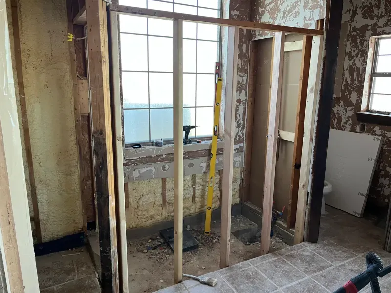 Interior view of a bathroom remodel. New wooden studs frame a shower, window visible.