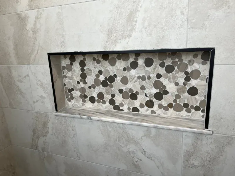 Recessed shower niche with pebble mosaic tile backing and stone shelf, surrounded by large neutral tiles.