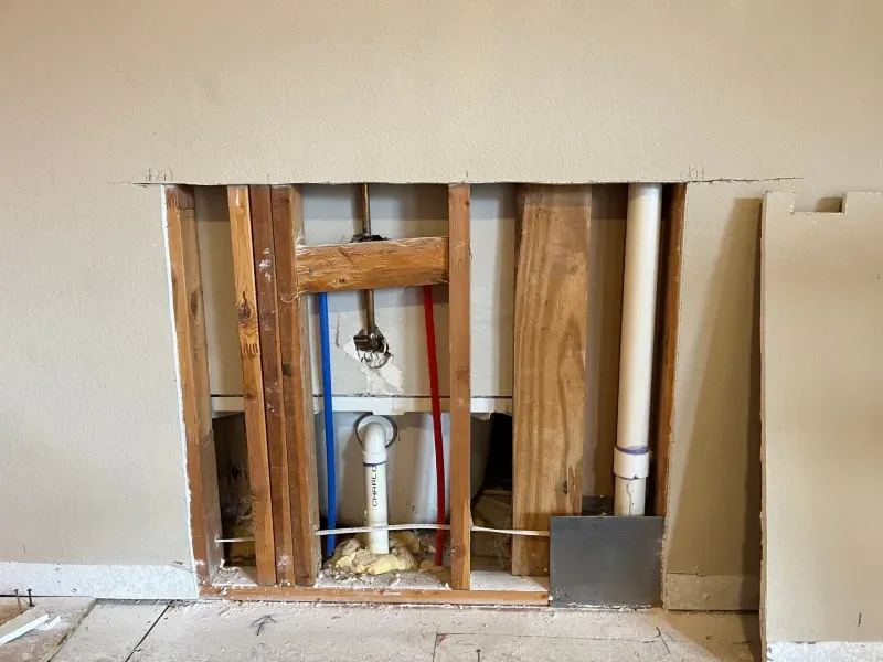 Wall opening revealing plumbing pipes and wooden framing. Blue, red, and white pipes are visible.
