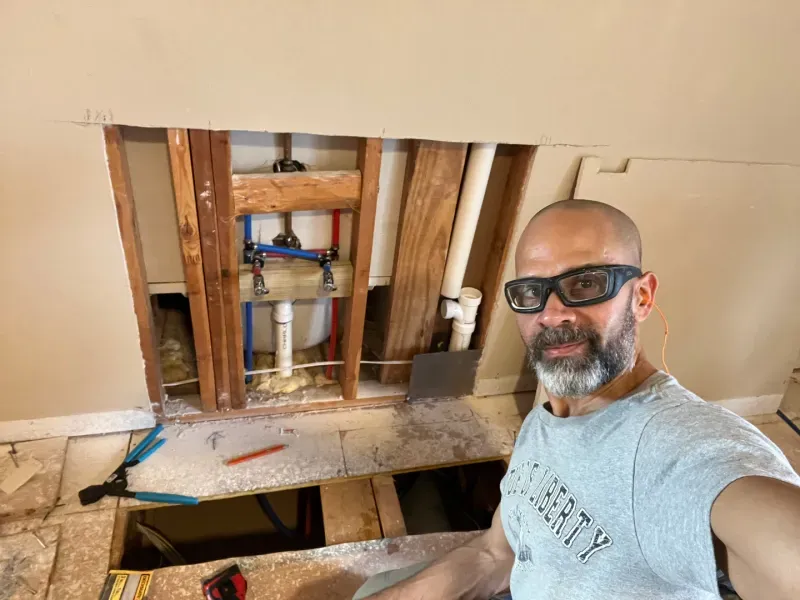 Man with safety glasses near open wall, plumbing visible. Construction setting.