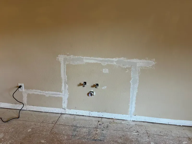 Wall with patched rectangular area, plumbing pipes, and electrical outlet; construction site setting.