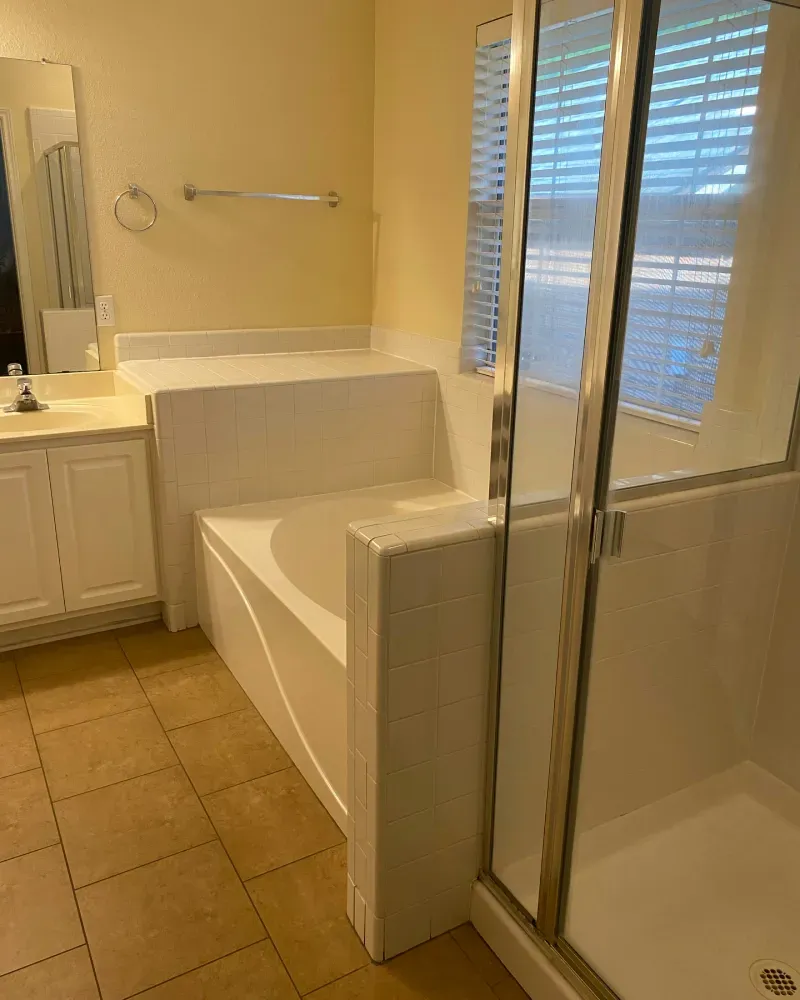 Bathroom with a built-in white bathtub, separate shower, cabinets, and a window with blinds.