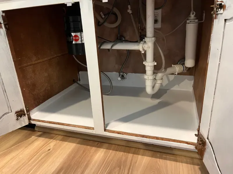 Inside of a white cabinet under a kitchen sink with plumbing, garbage disposal, and water filter.