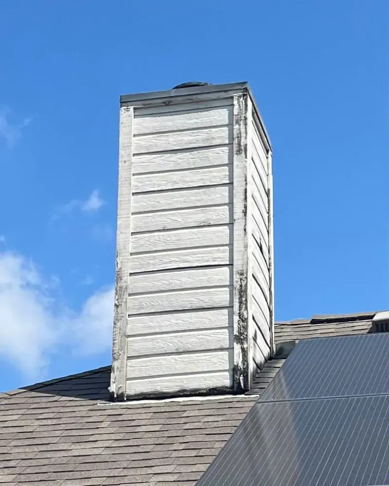 Chimney on a roof, white siding, blue sky, solar panels in foreground.