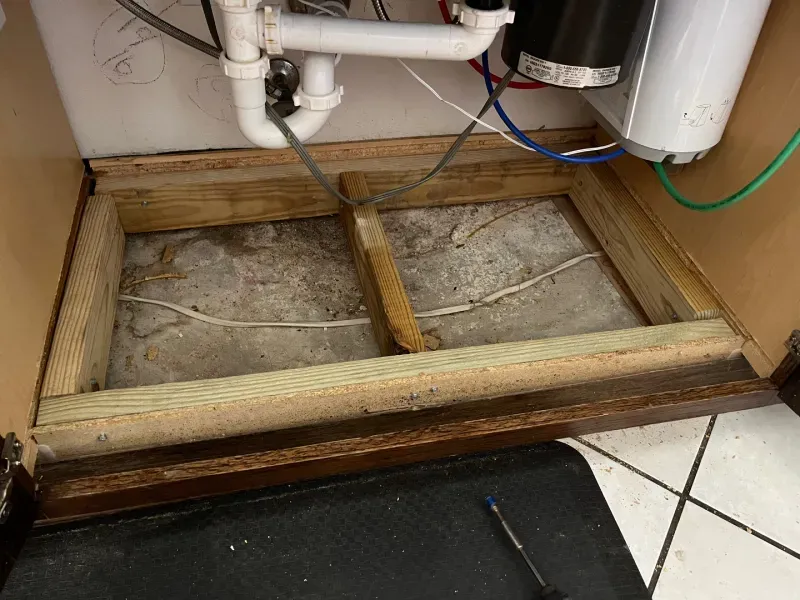 Wooden framework under a kitchen sink. Concrete floor visible. Plumbing, garbage disposal, and water filter are present.