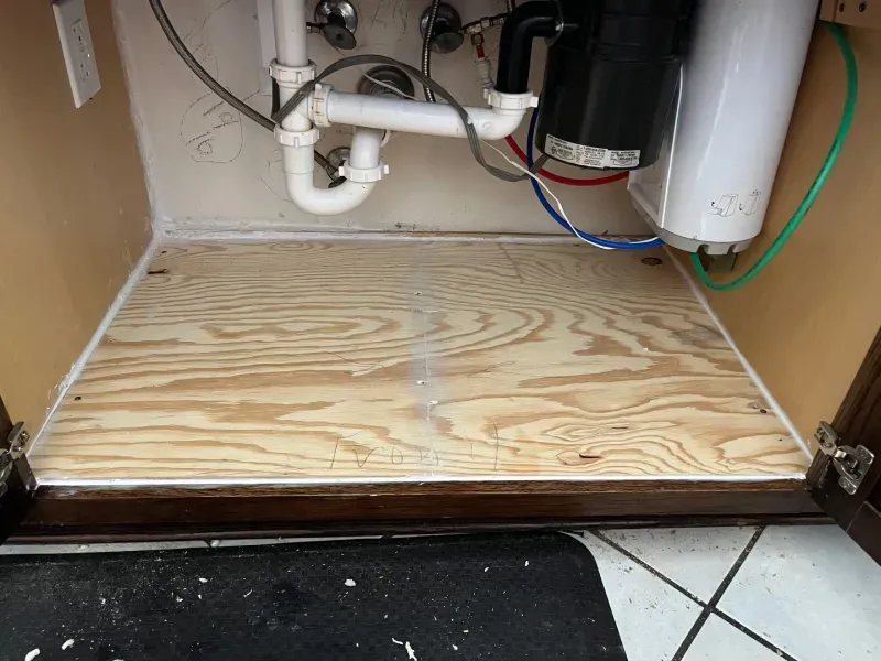 Inside of a kitchen cabinet with plywood base; plumbing, garbage disposal, water filter visible.