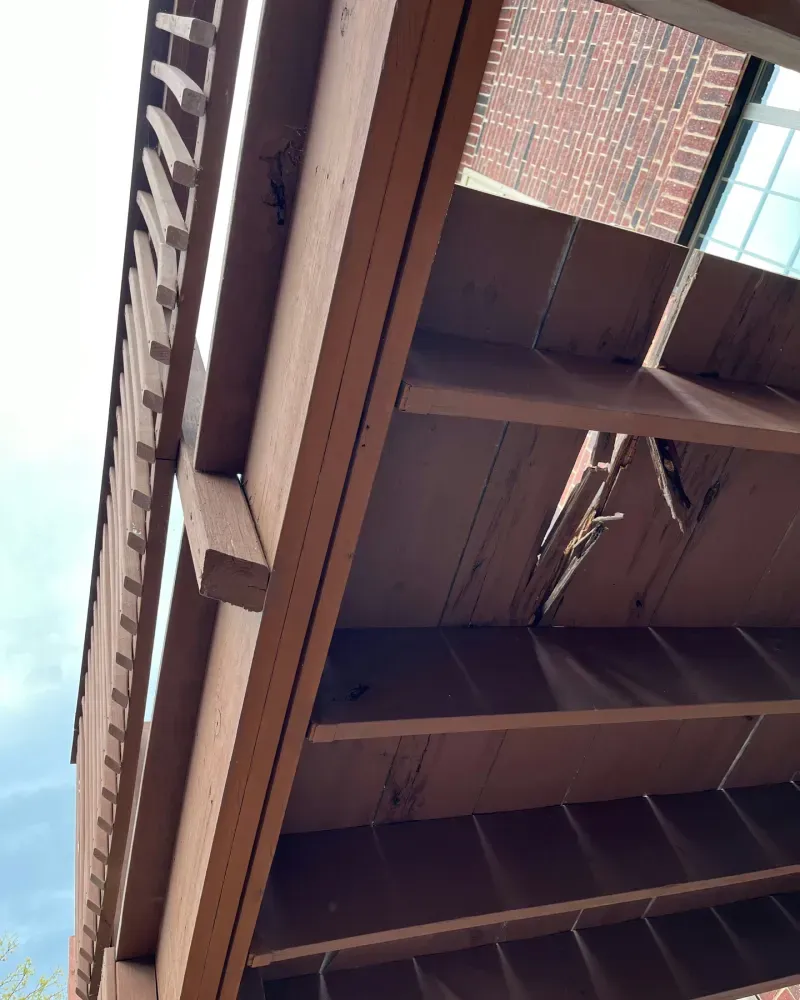 Low-angle view of a brown wooden pergola with damaged boards against a brick building and sky.