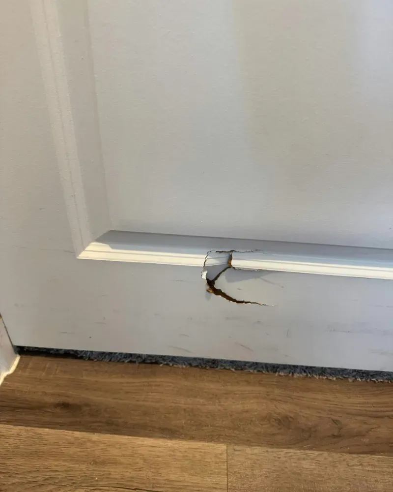 White trim on a wall with a crack near the floor. Brown wooden floor.