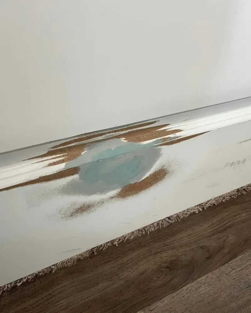Damaged white baseboard with turquoise paint over exposed wood, next to wood-look flooring.