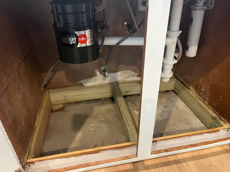 Under-sink cabinet with exposed plumbing, garbage disposal, and wooden support frame on the concrete floor.