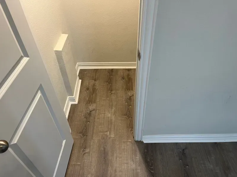Open doorway into a room with gray wood-look flooring and white trim, from a hallway with a partially open door.