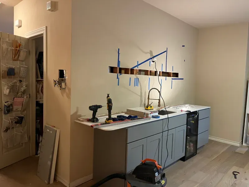 A kitchen renovation in progress. Wall with hole, blue cabinets, sink, tools on countertop, unfinished drywall.