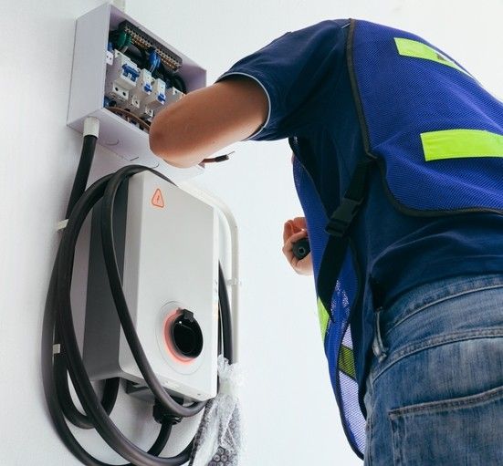Electrician installing an EV charger on a white wall. He's wearing a blue vest and working inside a junction box.