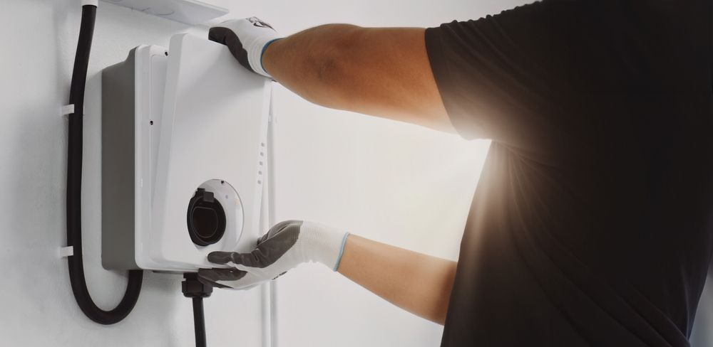 Person wearing gloves installing an electric vehicle charger on a white wall.