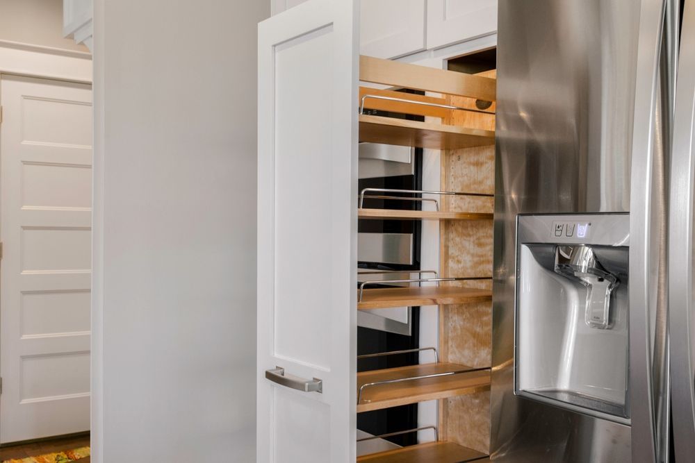 White kitchen pantry cabinet with pull-out shelves open, next to a stainless steel refrigerator with water dispenser.