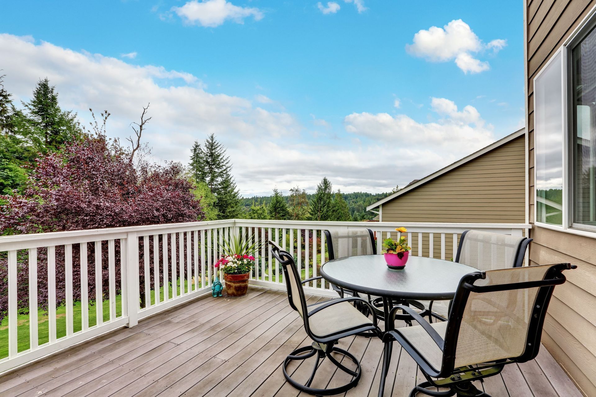 Deck with table and chairs, overlooking trees and a house, under a blue sky with clouds.