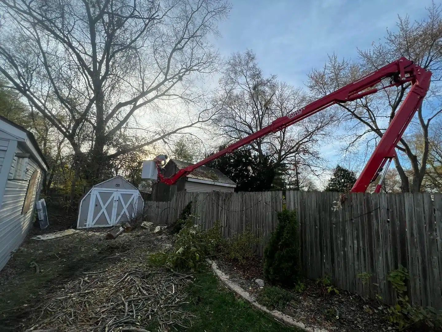Red concrete pump reaching over a wooden fence to pour concrete on a house roof, with a shed in the background.