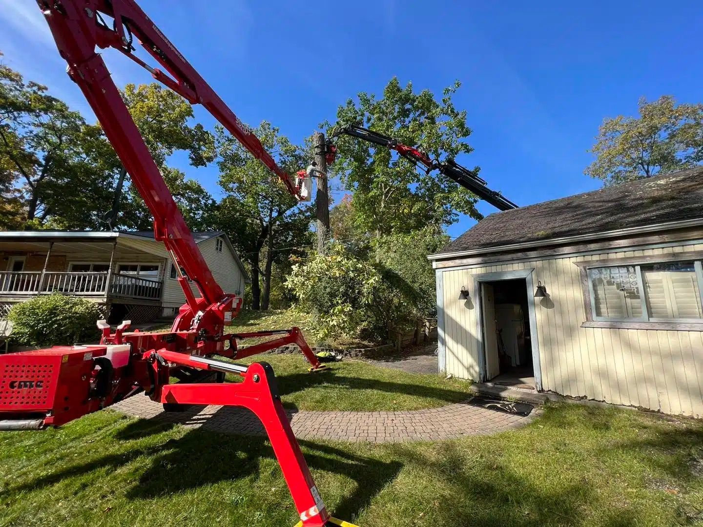 Red crane trimming a tree near a white building with a green lawn under a blue sky.