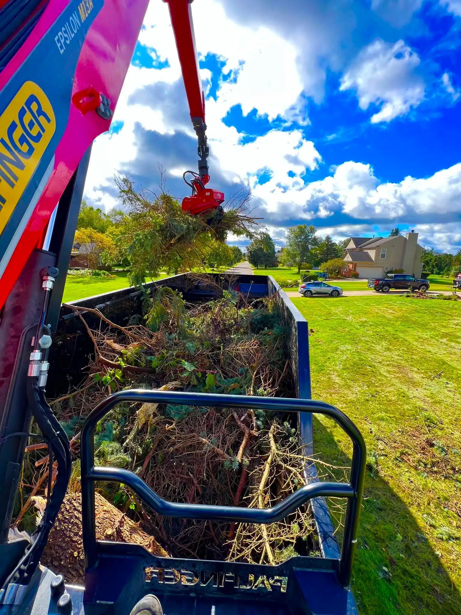A crane loading brush into a truck on a grassy lawn with a blue sky.