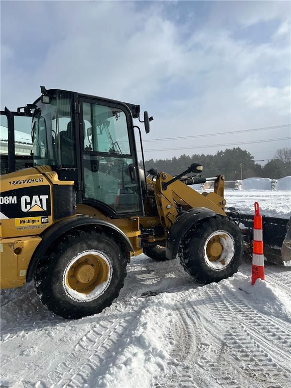 Yellow CAT loader on snow-covered ground with orange traffic cone. Cloudy day.