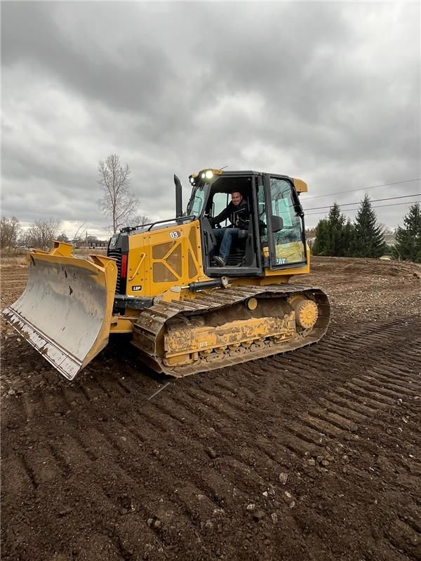 A person operates a yellow bulldozer on a dirt lot under a cloudy sky.