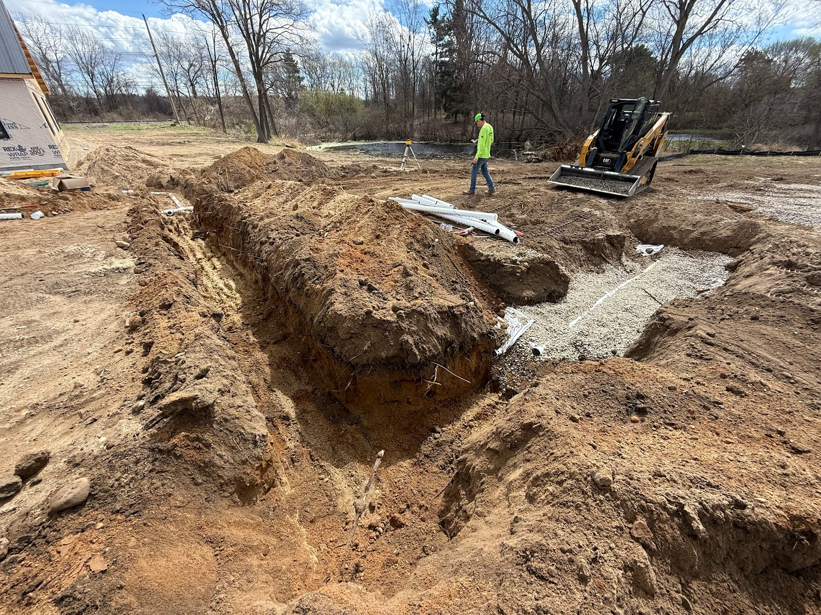 Construction site: trenches dug in dirt, person in yellow vest, small excavator, cloudy sky.
