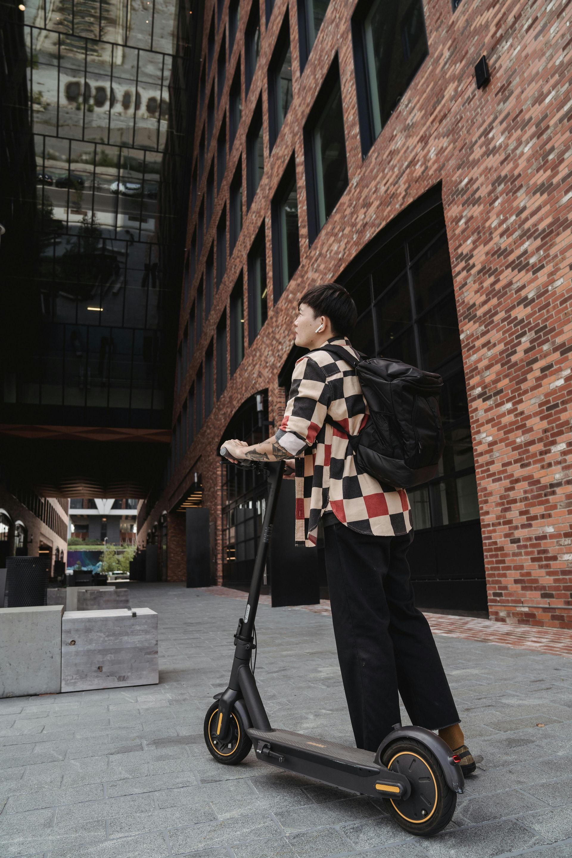 Person on scooter in front of a brick building, wearing checkered shirt and backpack. — Freedom Sports in Bundaberg Central, QLD