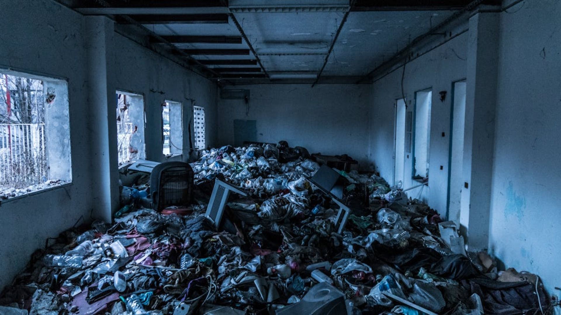 Interior de una habitación descuidado, lleno de basura y escombros; se ven ventanas y puertas.
