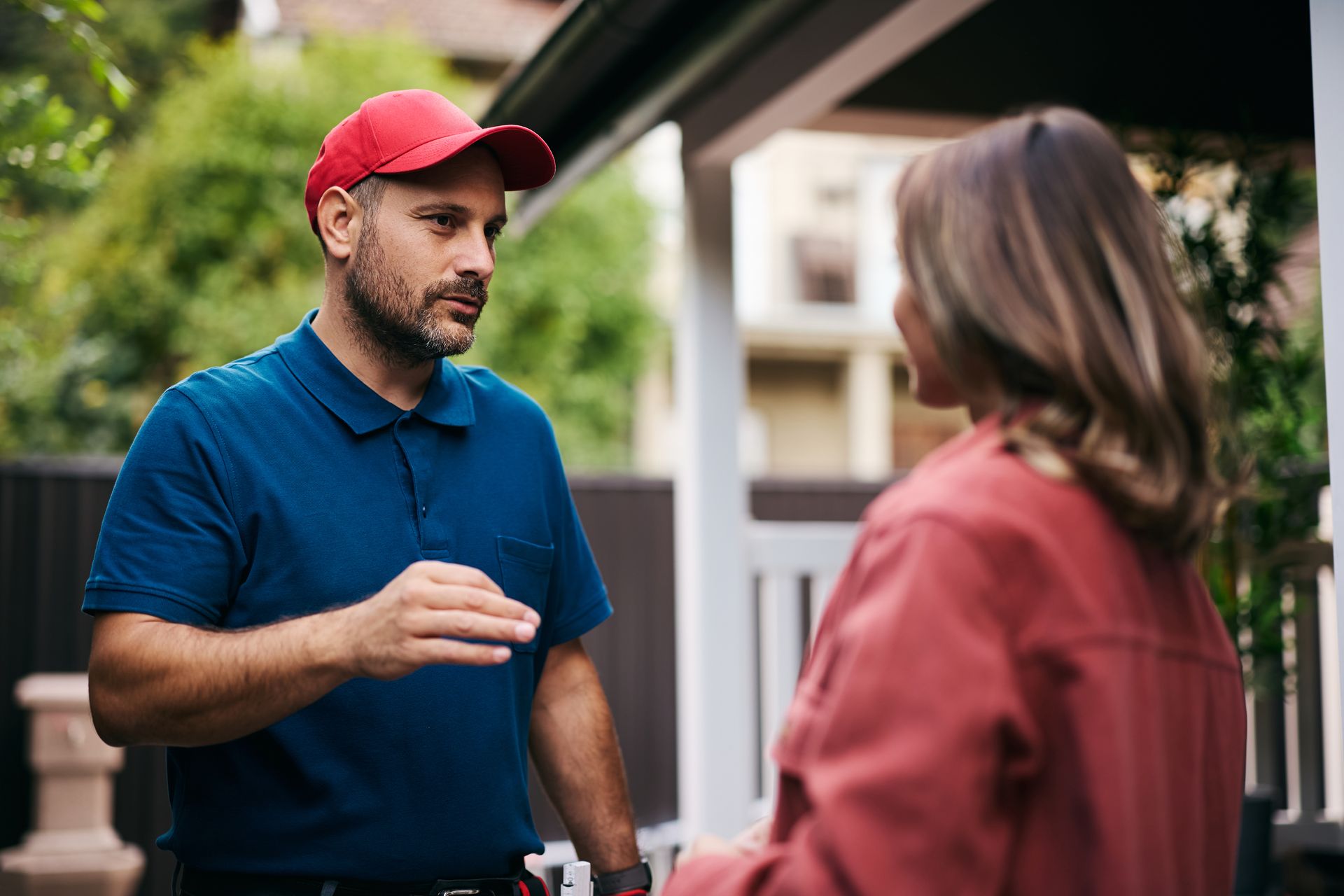 Two people talking outside a house, one in a red cap and blue shirt, the other in a pink jacket.