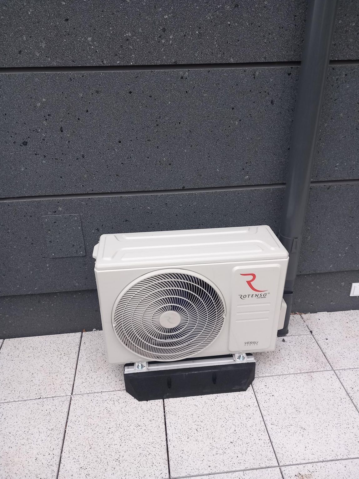 Jednostka zewnętrzna klimatyzatora zamontowana na stopach bitumicznych, zapewniających stabilność i pochłanianie drgań.