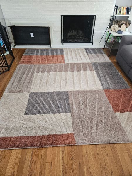 Geometric patterned rug with gray, cream, and rust-colored sections on a wooden floor in front of a fireplace.