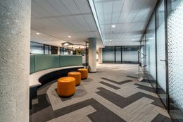 Modern office waiting area with curved teal bench, orange stools, patterned gray carpet, glass walls.