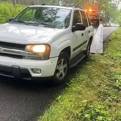 White SUV being towed on a narrow road with a tow truck and flashing lights.