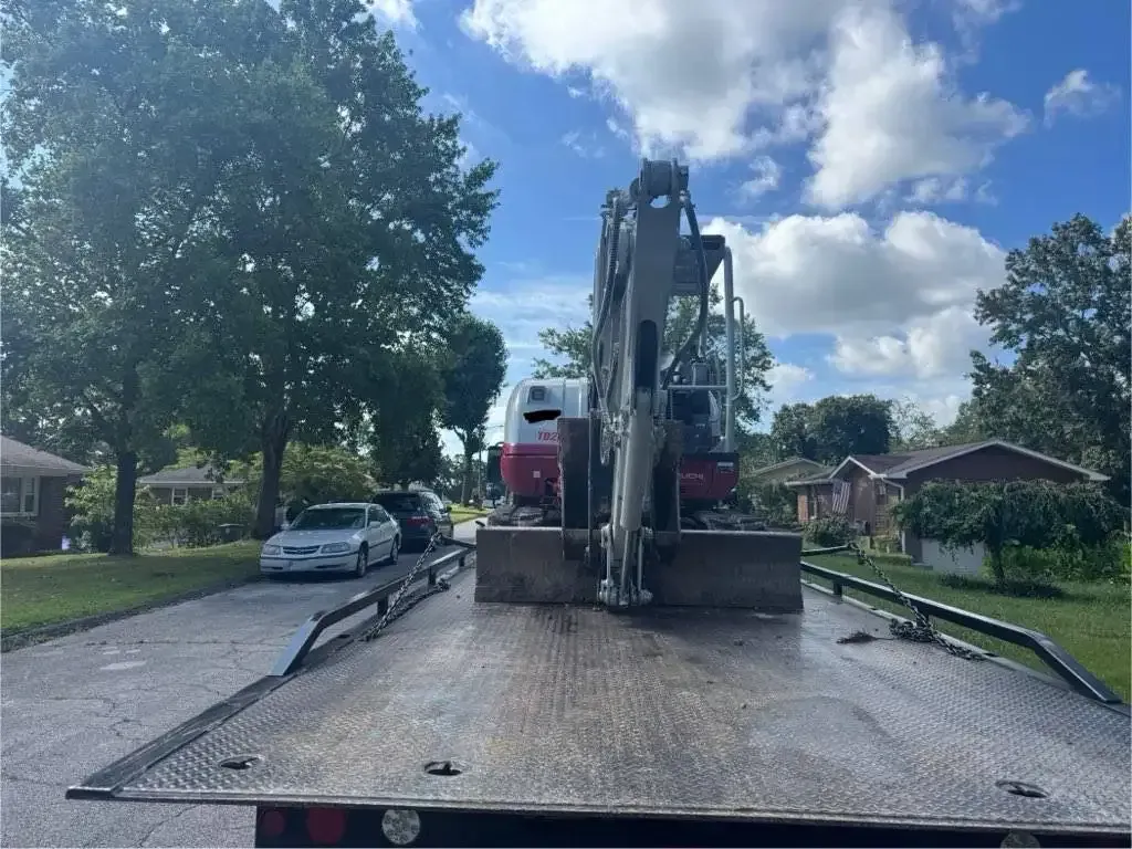 A mini excavator on a flatbed tow truck on a residential street under a blue, cloudy sky.