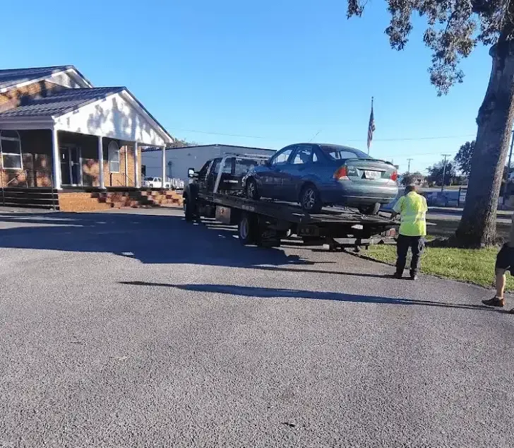 Tow truck with a blue car on its bed, parked outside a building on a sunny day. A person stands nearby.