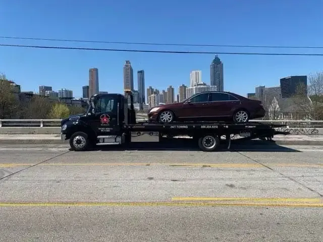 Tow truck carrying a maroon sedan on a city bridge, Atlanta skyline in background.