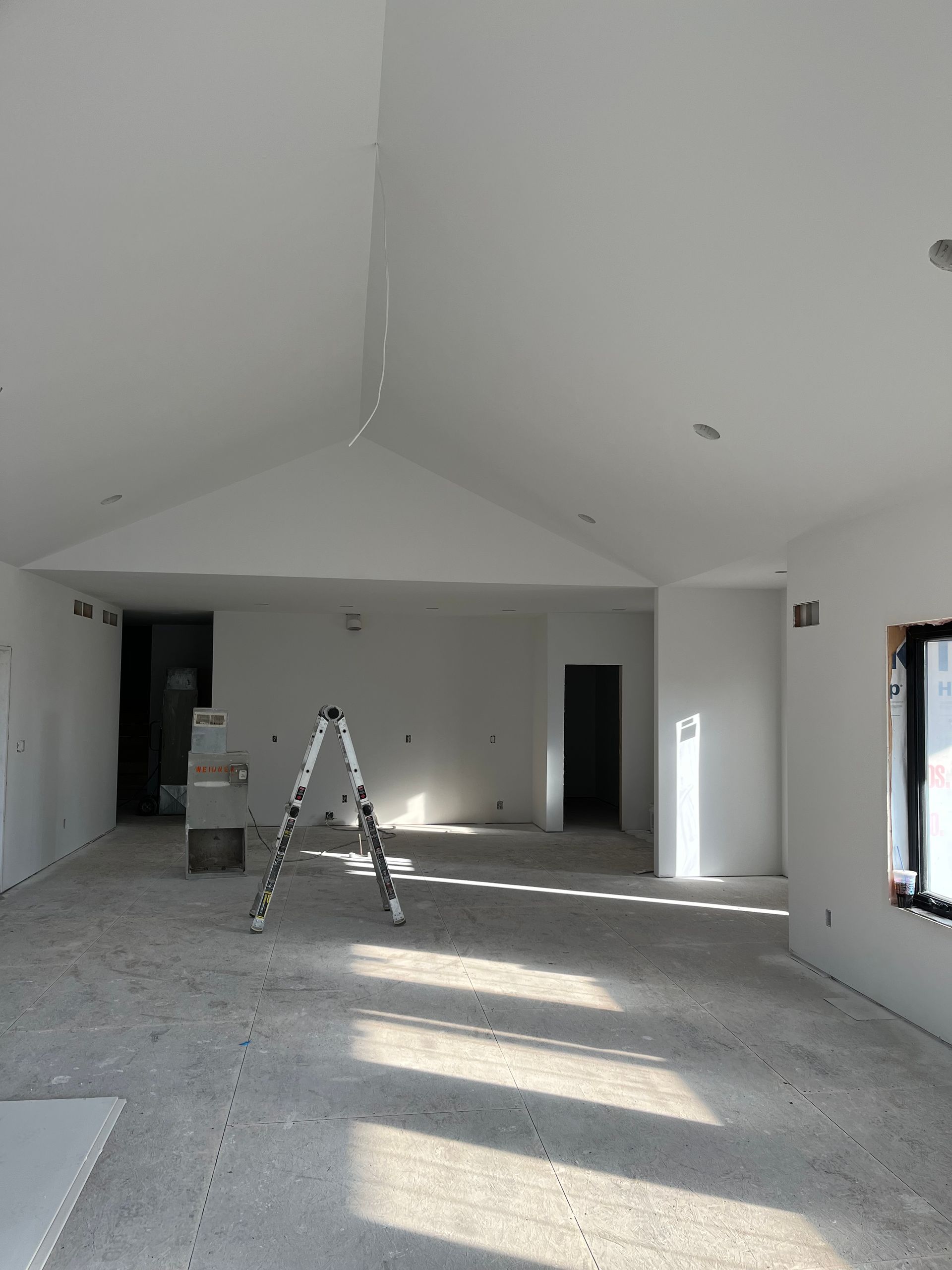 Interior of a room under construction with white walls, a high ceiling, and a ladder in the center.
