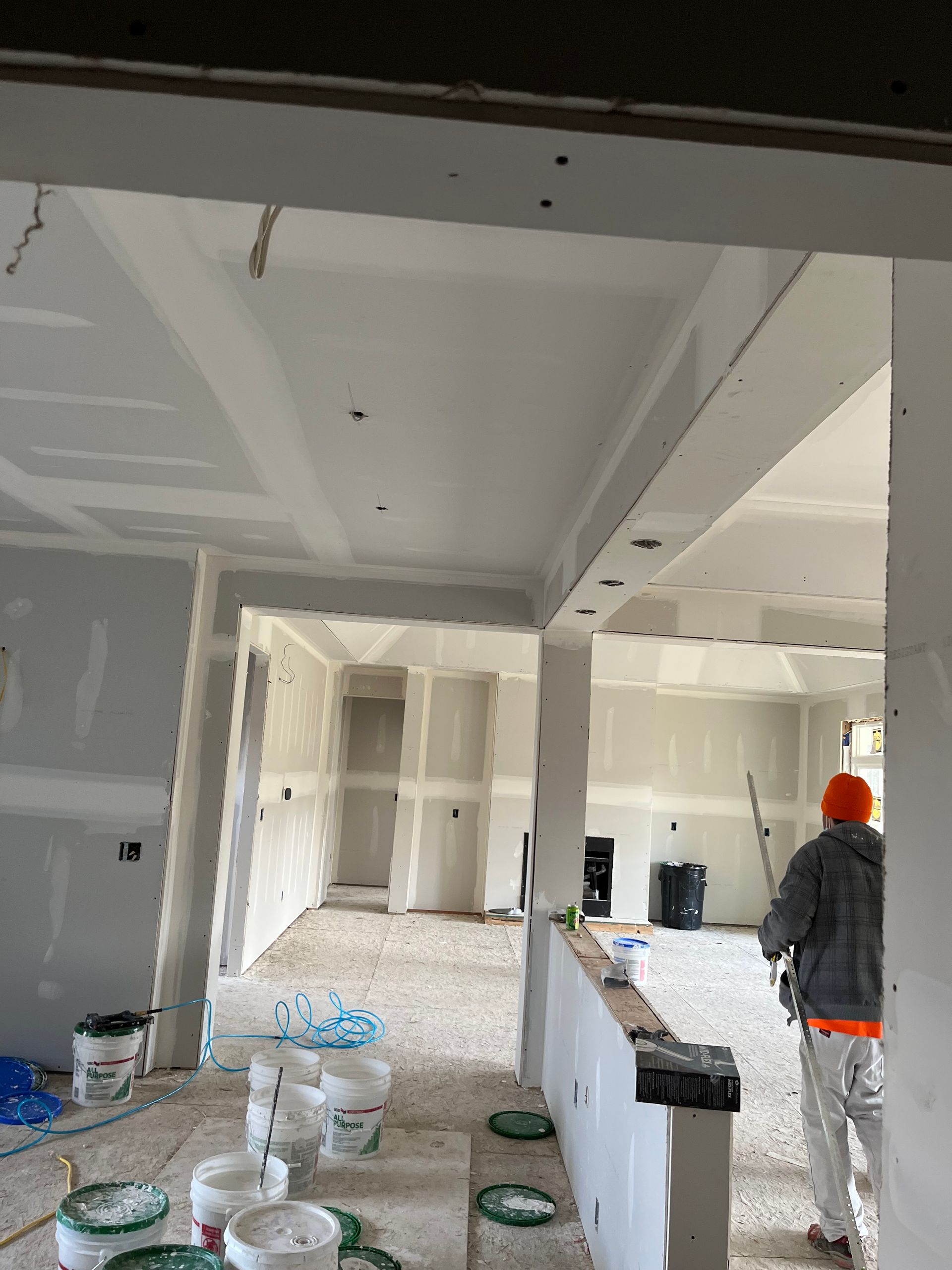 Interior construction site with drywall. A worker stands near a partially built wall. Buckets and debris are visible.