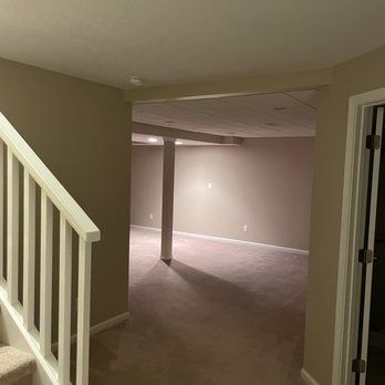 Interior view of a basement hallway with stairs, doorway, and an open room with carpet. Beige walls.