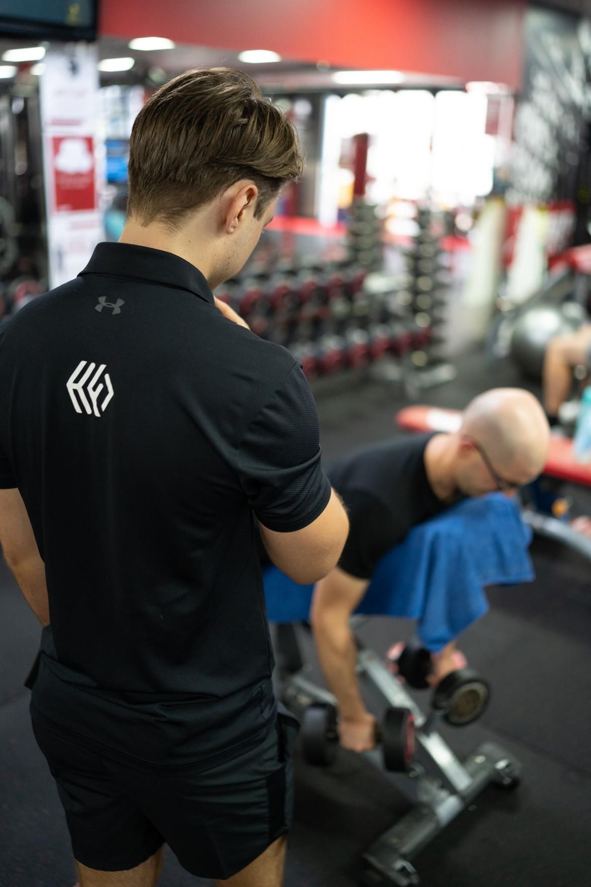 A Man is Standing Next to a Man Lifting Dumbbells in a Gym — Health First Townsville In Townsville, QLD
