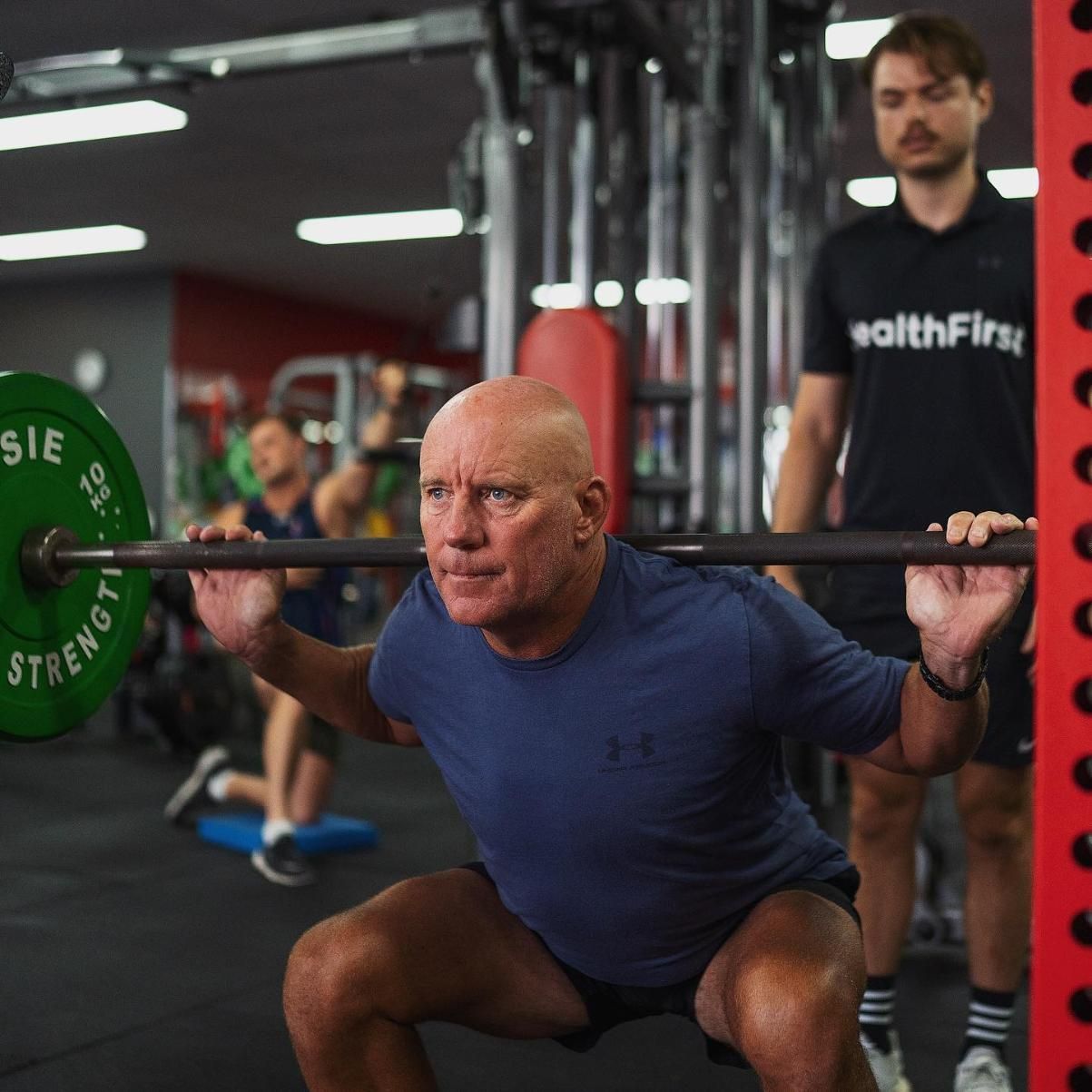 A Man is Squatting With a Barbell in a Gym β Health First Townsville In Townsville, QLD