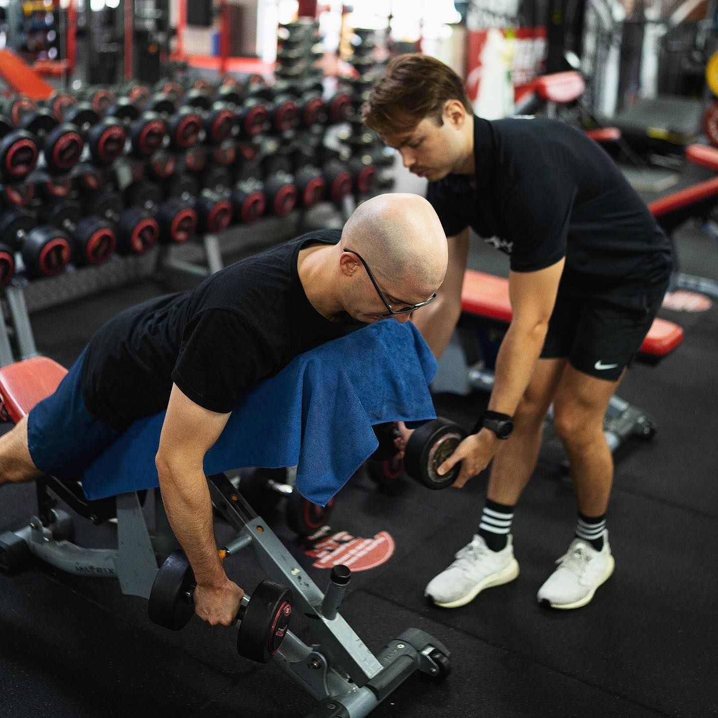A Man is Helping Another Man Lift a Dumbbell in a Gym β Health First Townsville In Townsville, QLD