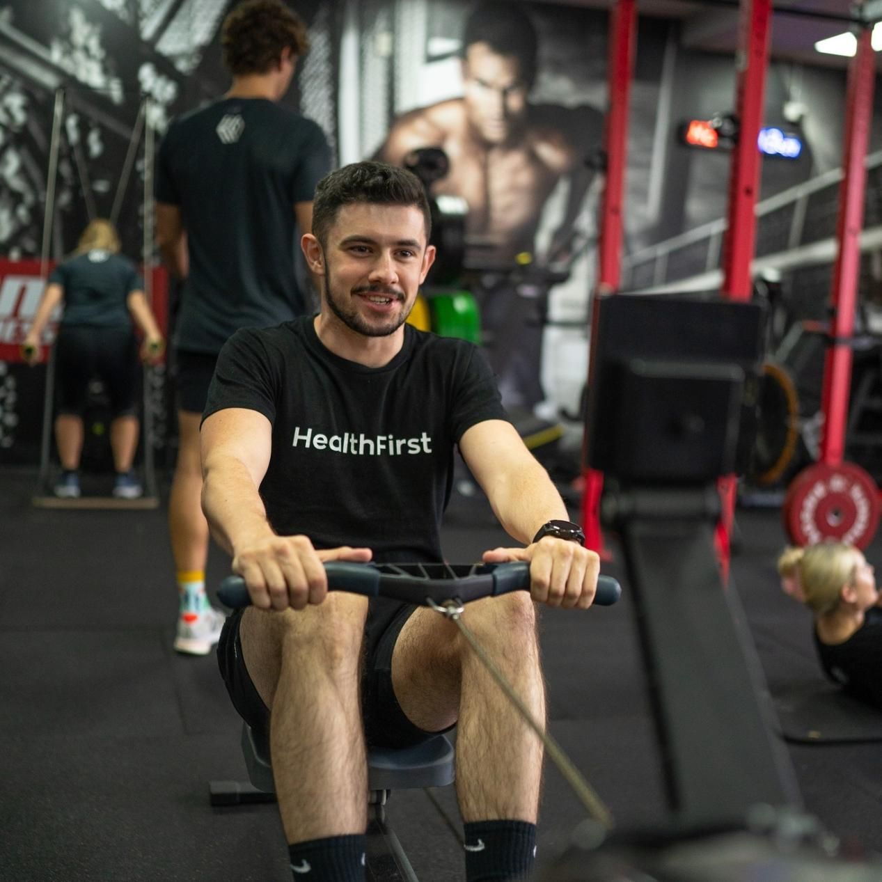 A Man Wearing a Black Shirt That Says Health First is Rowing in a Gym β Health First Townsville In Townsville, QLD