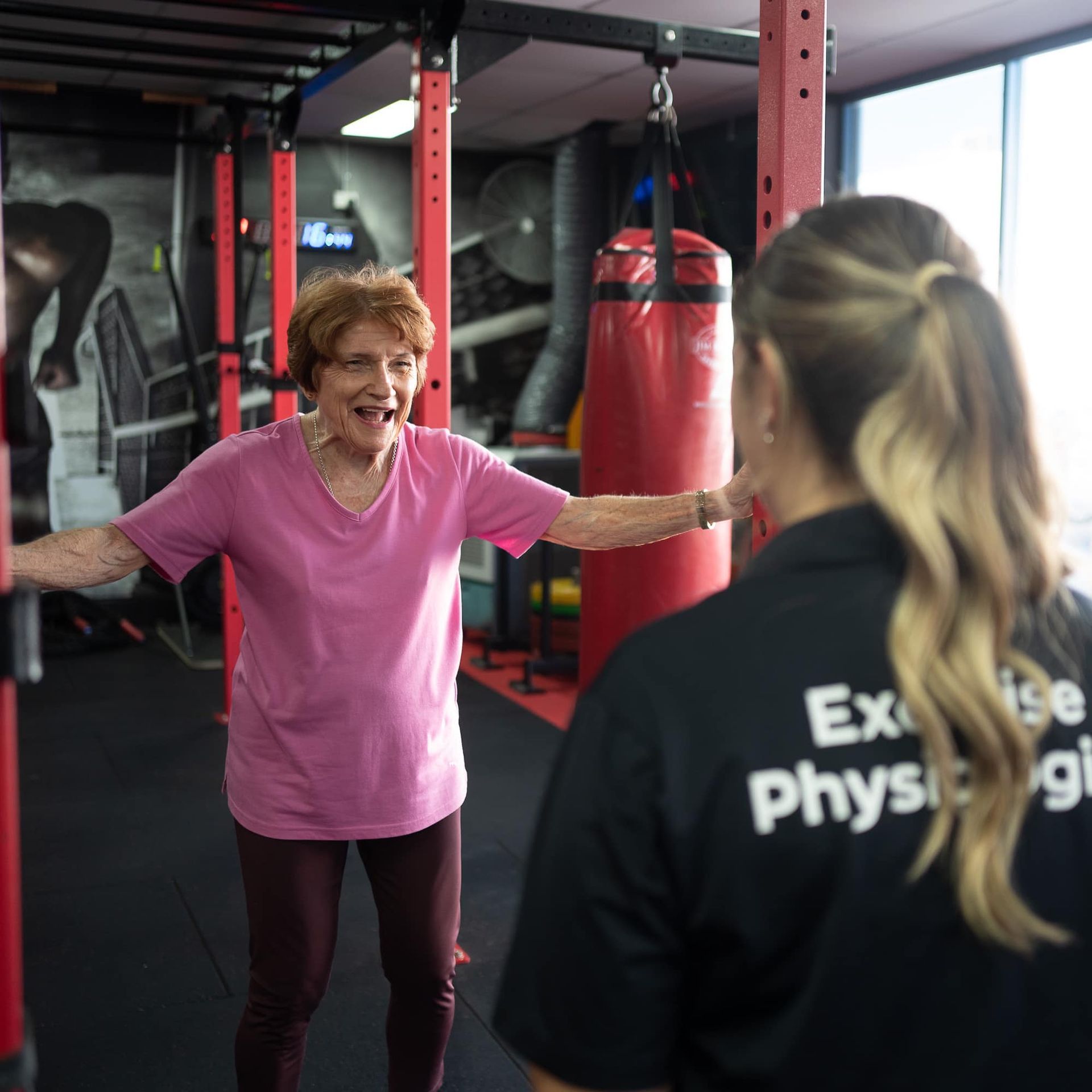 Older woman in pink shirt smiles, arms out, in gym. Trainer watches.