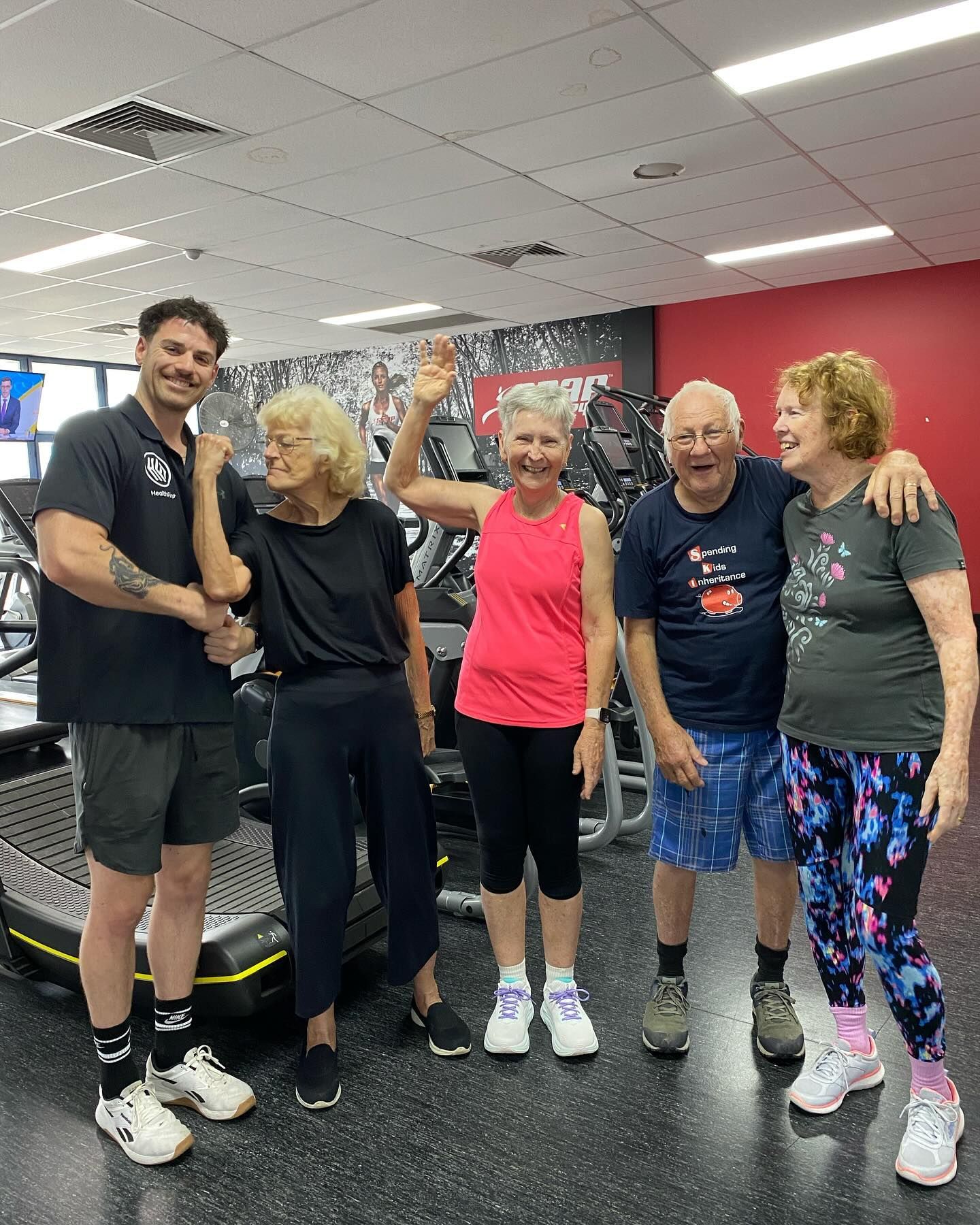 Man squatting with barbell in gym, spotter behind, other people exercising — Health First Townsville In Townsville, QLD