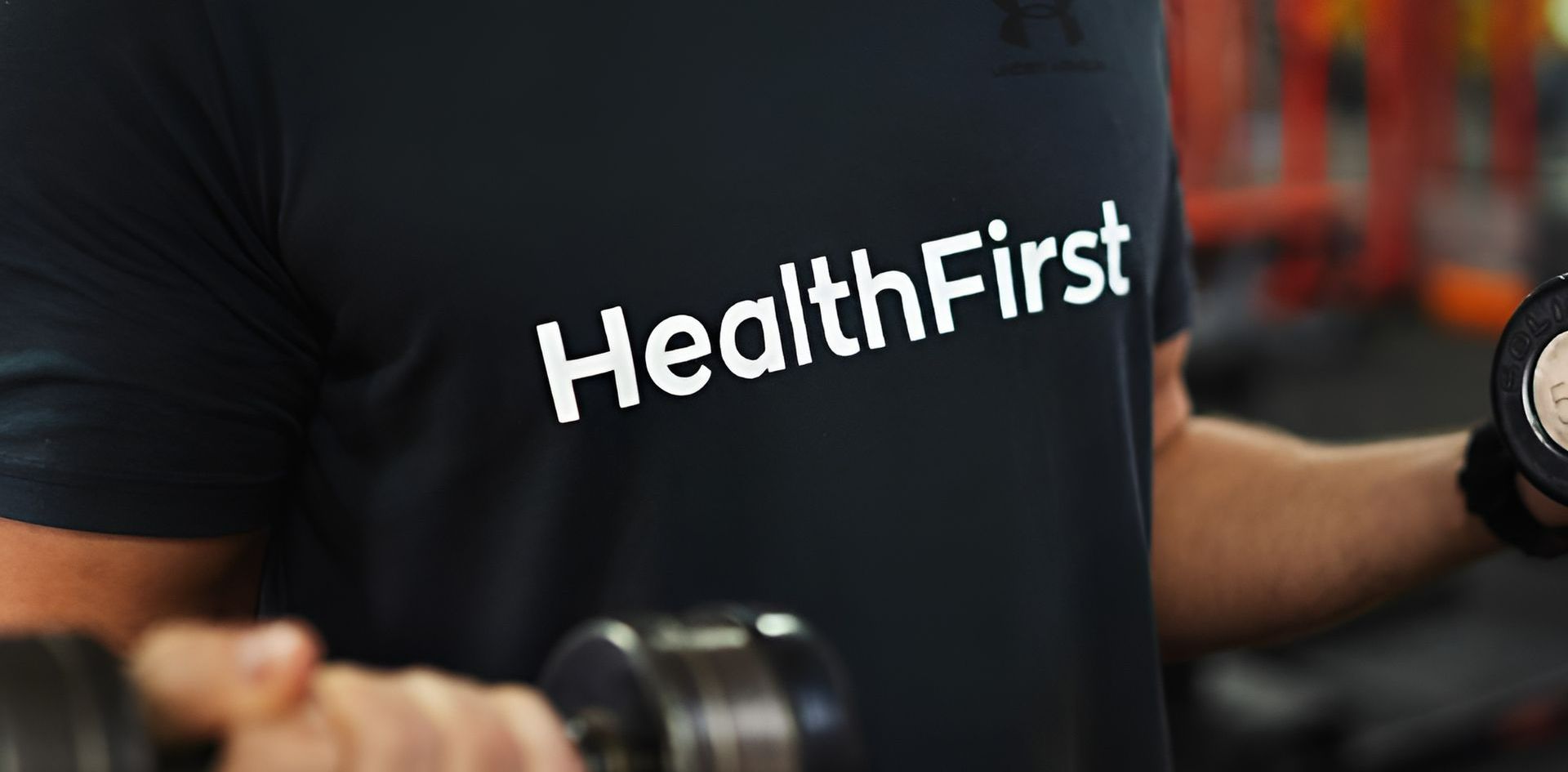 Man in Black Shirt With "Healthfirst" Logo, Lifting Weights in a Gym — Health First Townsville In Townsville, QLD