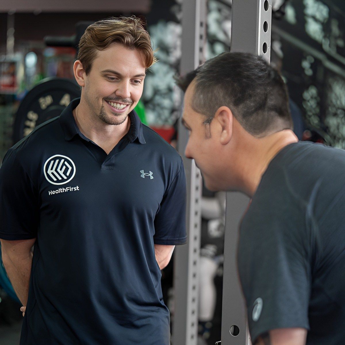 A Man in a Black Under Armour Shirt is Talking to Another Man in a Gym — Health First Townsville In Townsville, QLD