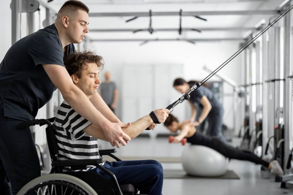 A Man in A Wheelchair Is Being Helped by A Man in A Gym — AMC Exercise Physiology in Maitland, NSW