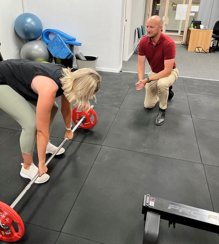 A Man is Watching a Woman Lift a Barbell in a Gym — AMC Exercise Physiology in Maitland, NSW