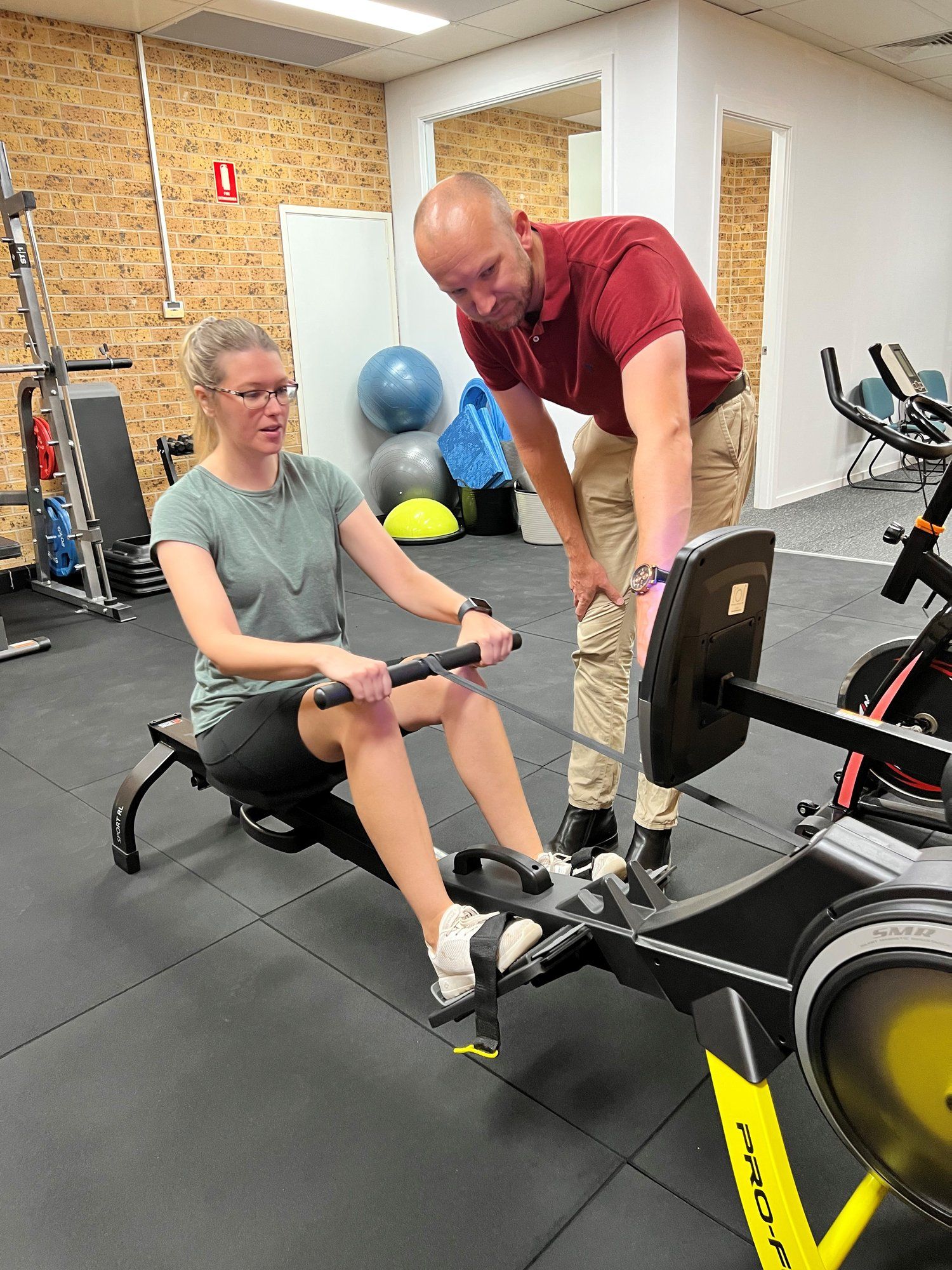 A Man Is Teaching a Woman how To Use a Rowing Machine in A Gym — AMC Exercise Physiology in Maitland, NSW